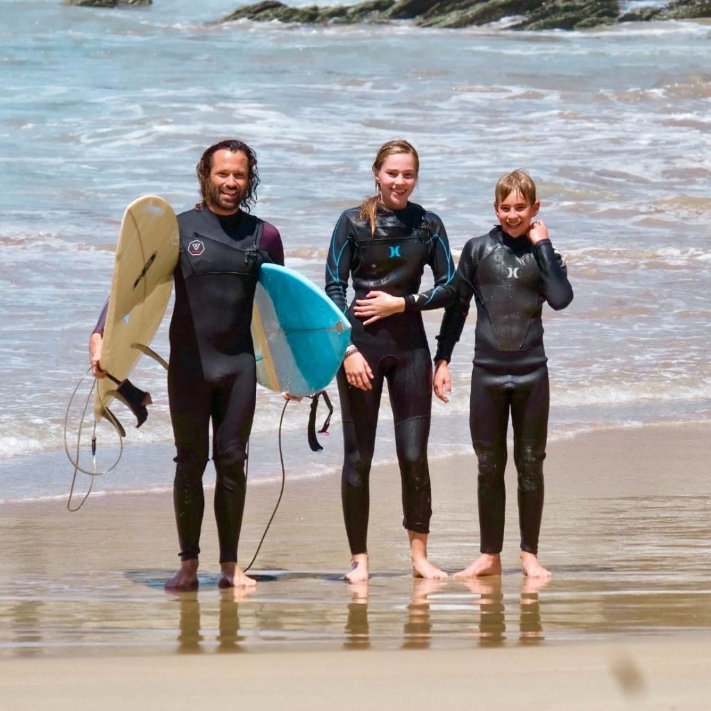 An adult male and two teenagers standing in wetsuits on the shore holding surfboards.