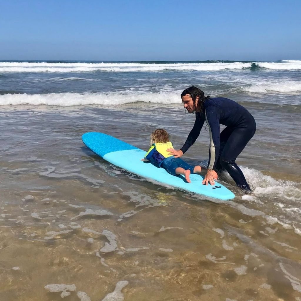 A surf instructor in a wetsuit assists a child lying on a blue surfboard in shallow ocean water.