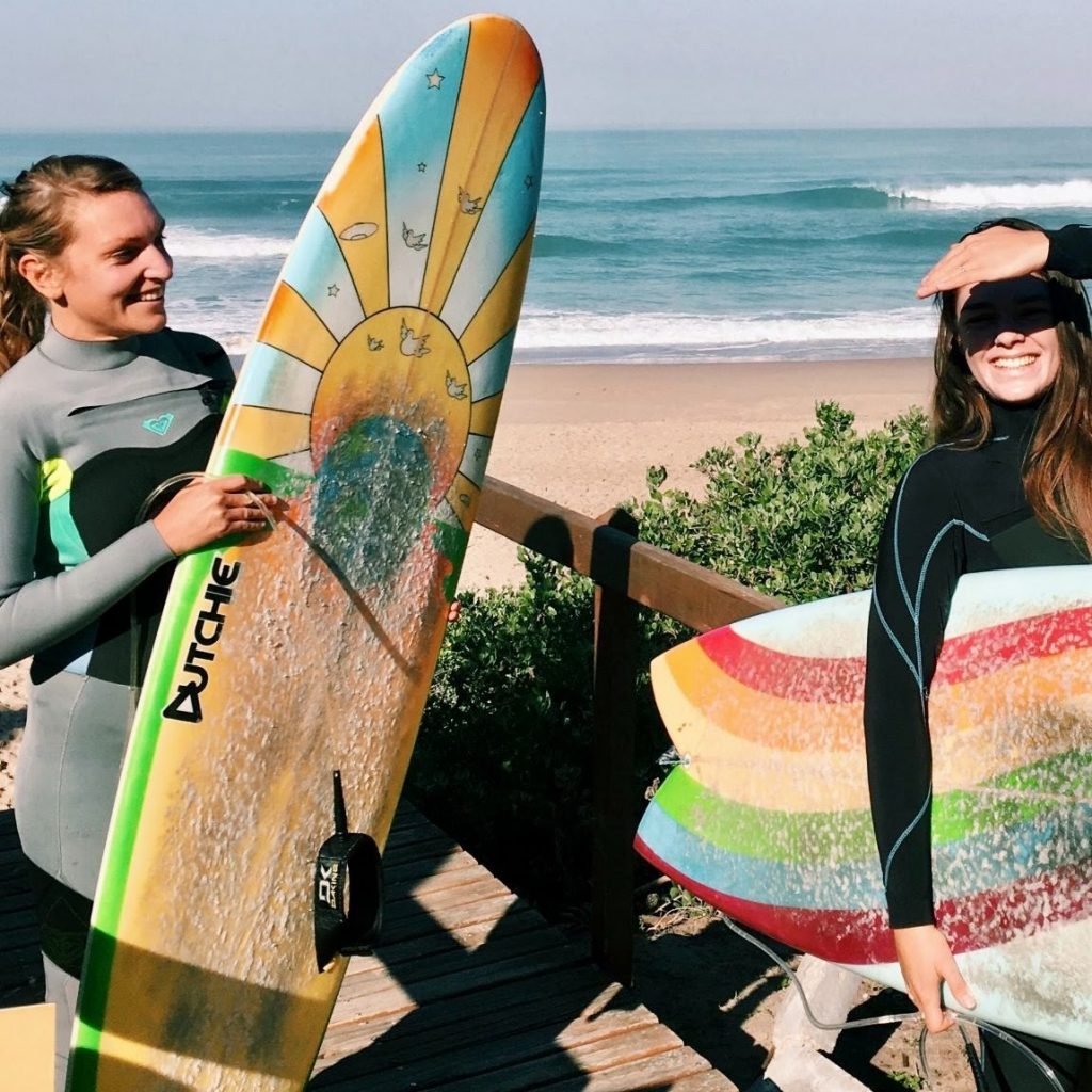 Two women in wetsuits holding surfboards on a wooden walkway with a sunny beach and ocean in the background.