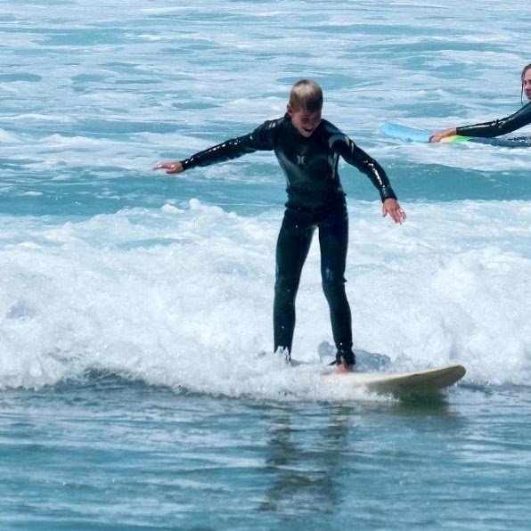 Young boy in a black wetsuit balancing on a surfboard while riding a small wave in the ocean.