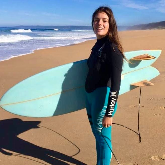 A smiling young woman in a black and teal wetsuit holding a light blue surfboard while standing on a sandy beach.