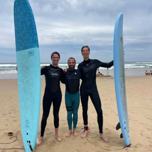 Three men in wetsuits standing on a sandy beach, holding surfboards on either side.