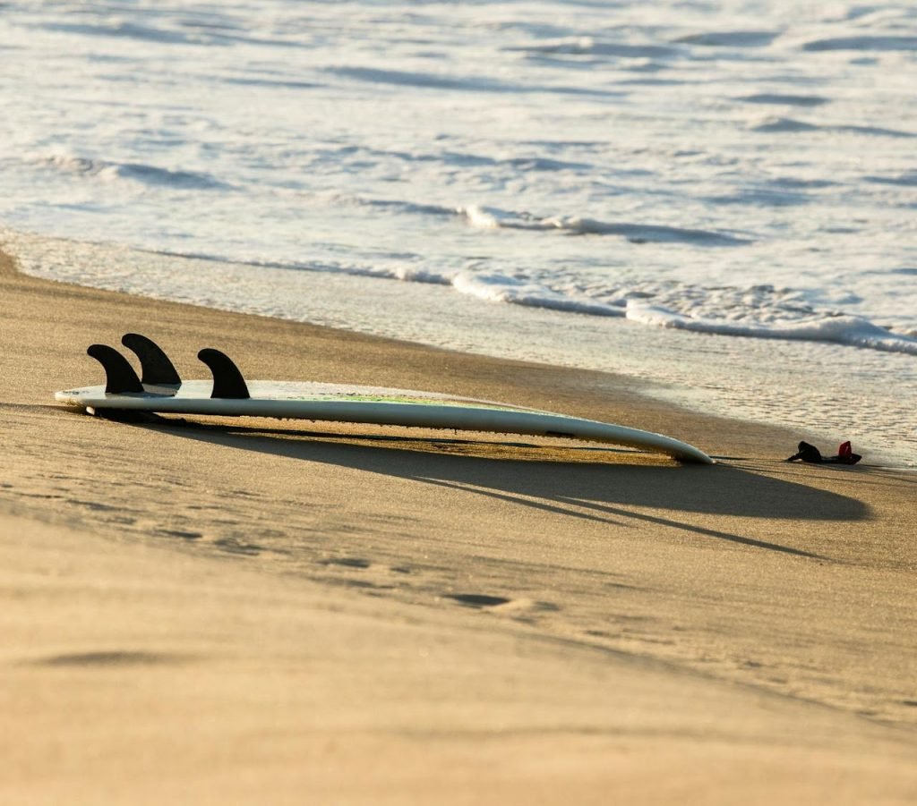 Surfboard with three black fins resting on a sandy beach near the shoreline with light foam from waves.