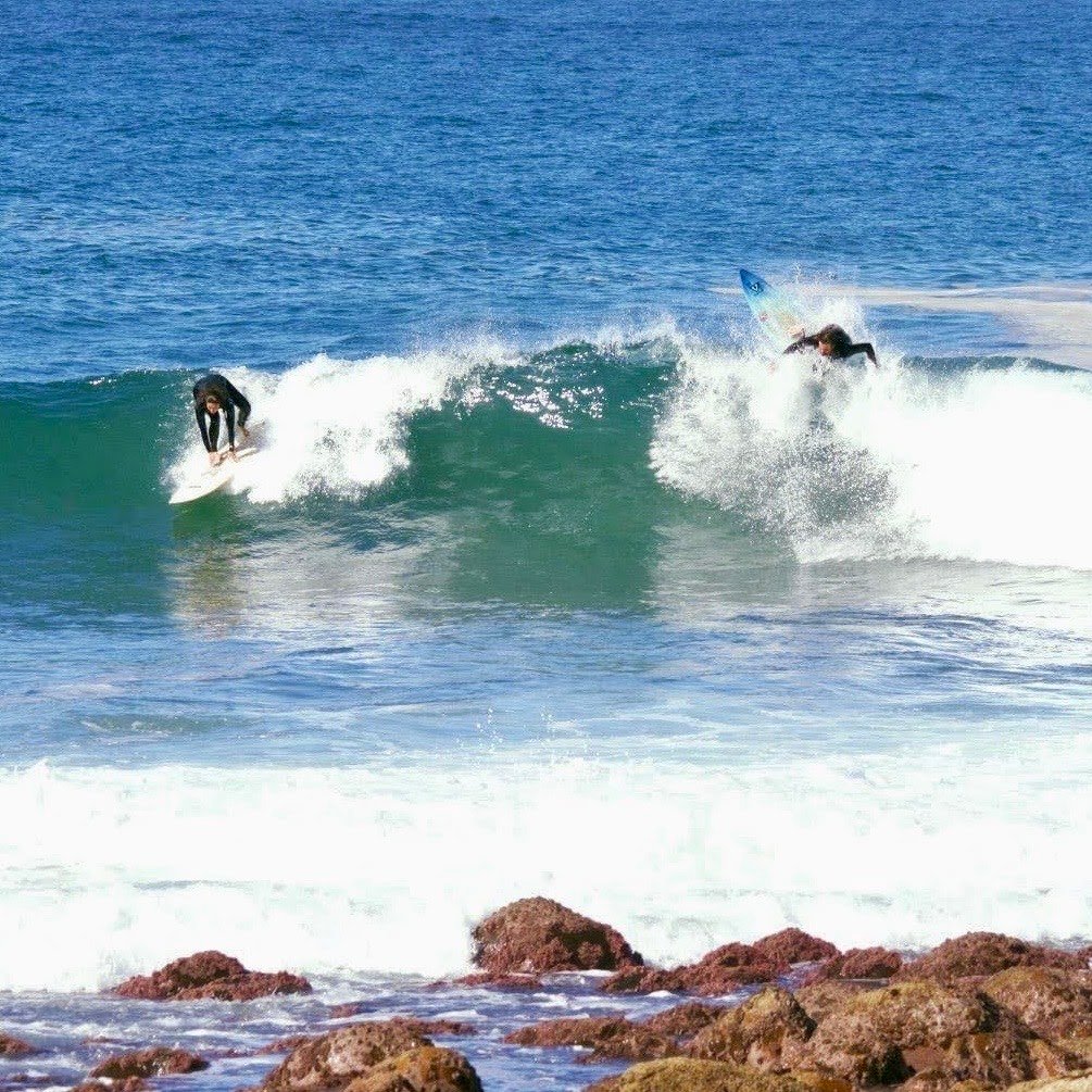 Two surfers in black wetsuits riding waves near a rocky shoreline.