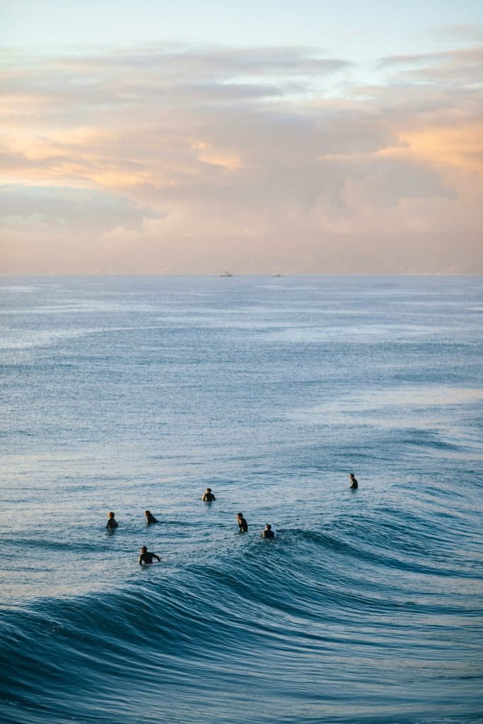 Seven surfers in black wetsuits waiting for waves in the ocean during a soft, overcast sunrise.