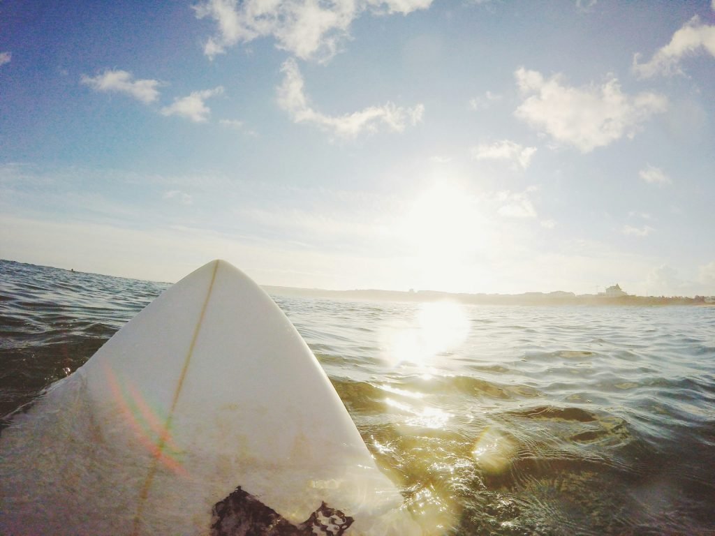 POV view from the nose of a white surfboard resting on calm ocean water toward a bright sun on the horizon.